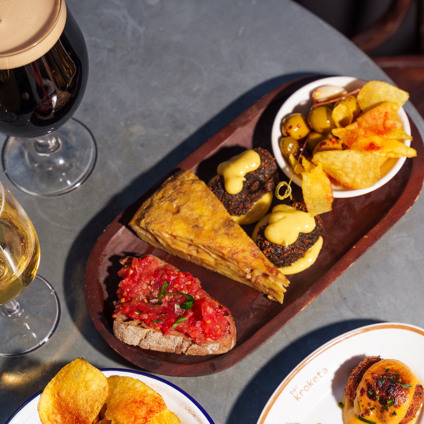 A wooden board with a variety of food including tortilla, olives, crisps and pan con tomate.