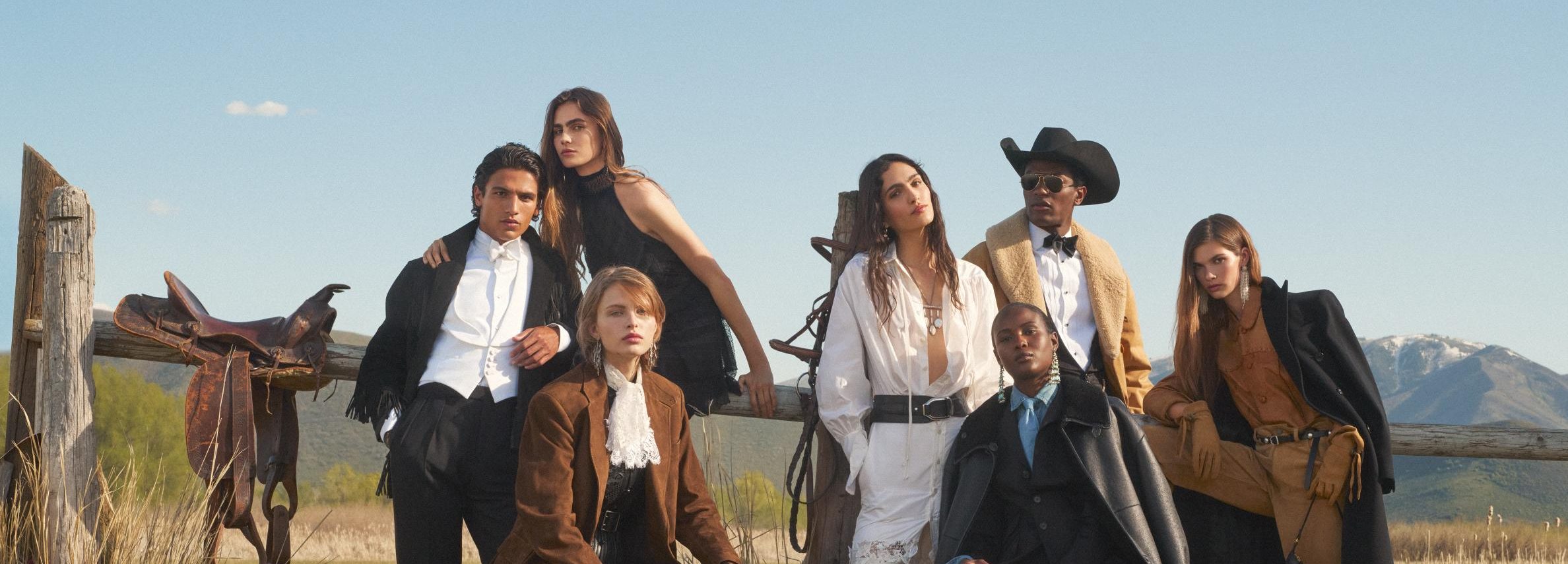 The photo features a group of seven models posing outdoors in what appears to be a Western or ranch setting, possibly in the mountains, given the background. They are dressed in a mix of elegant, rugged, and Western-inspired clothing, which is a signature style for the brand.