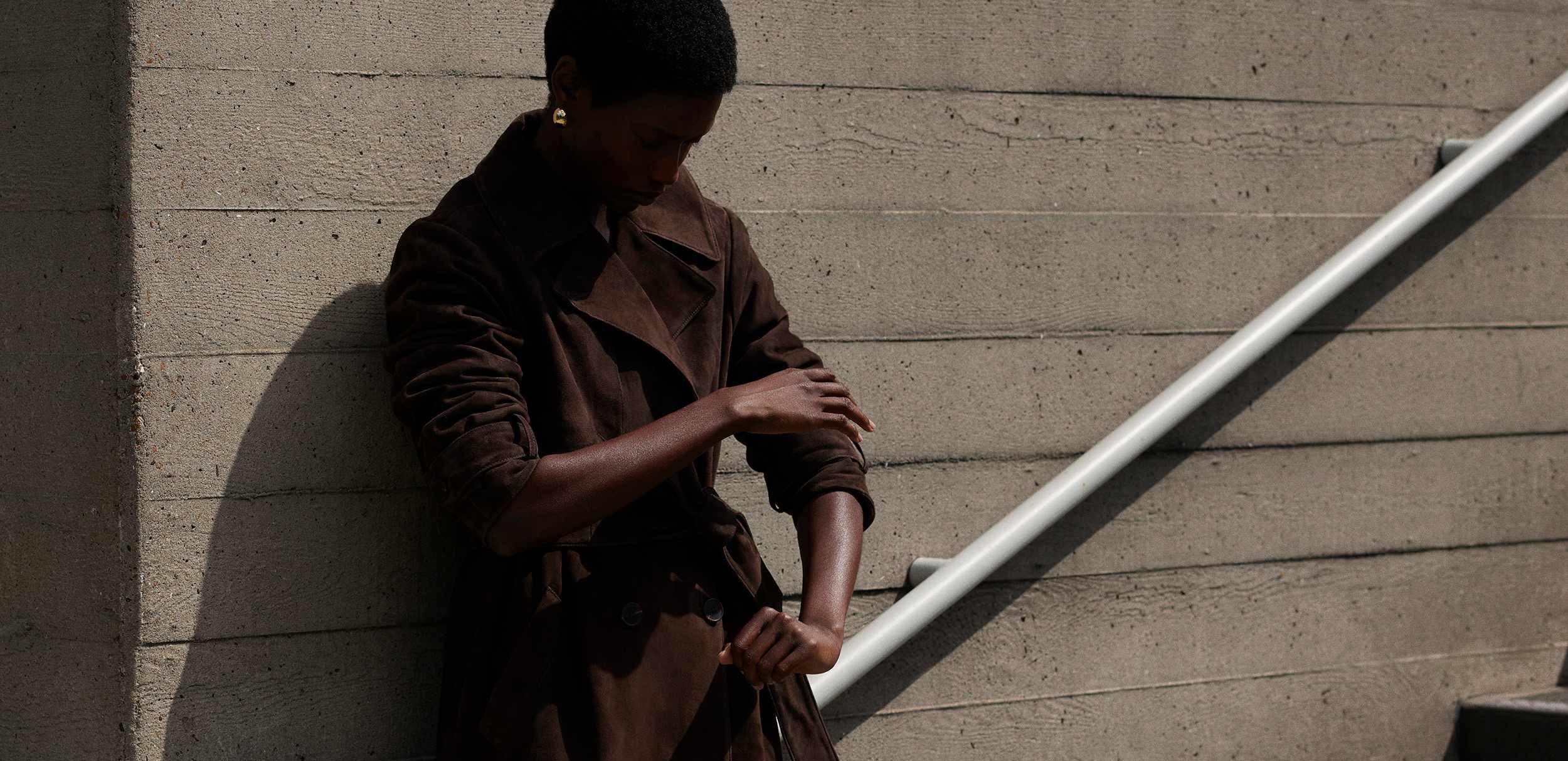 outdoor fashion shot featuring a person wearing a long, dark brown coat that looks like it could be suede or another soft material, styled with a tie-belt.