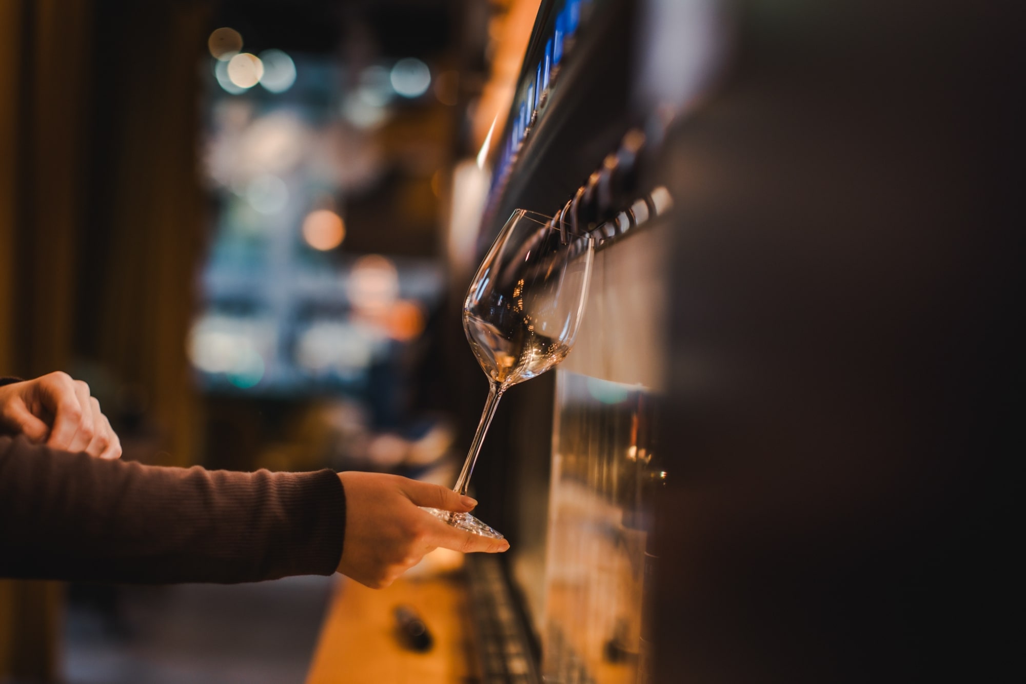 The image captures a close-up, somewhat dimly lit scene in what appears to be a bar, wine cellar, or tasting room. The focus is on a delicate, empty wine glass being held by a person's hand and forearm. The person's sleeve is a dark, ribbed fabric, possibly brown or burgundy.