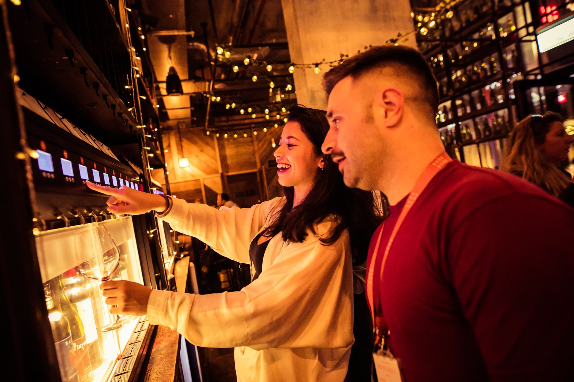 The image captures a close-up, candid moment of a man and a woman interacting with a modern wine dispensing machine in a dimly lit, warm-toned bar or tasting room. The scene is full of energy and engagement.