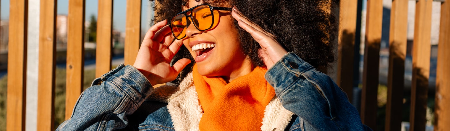 he image shows a person from the chest up, captured outdoors on a sunny day. They are smiling widely with a joyful expression and are adjusting a pair of sunglasses with bright orange-tinted lenses.
