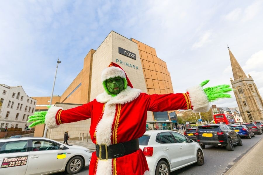 In the foreground, a person dressed as the Grinch is standing with their arms outstretched, giving a welcoming or dramatic gesture. They are wearing a Santa Claus suit with white fur trim, a matching hat, and a wide black belt. The person has bright green face makeup and shaggy green gloves extending past their elbows.