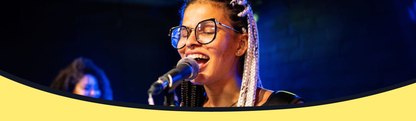 **Alt text:** A close-up shot of a female lead singer with glasses and braided hair singing into a microphone on a dimly lit stage. In the background, a guitarist is softly blurred under cool blue stage lighting.