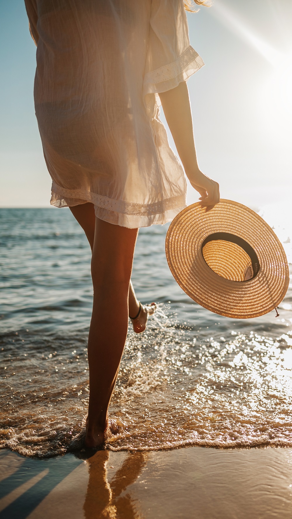 A woman walks along a sunny beach holding a sun hat