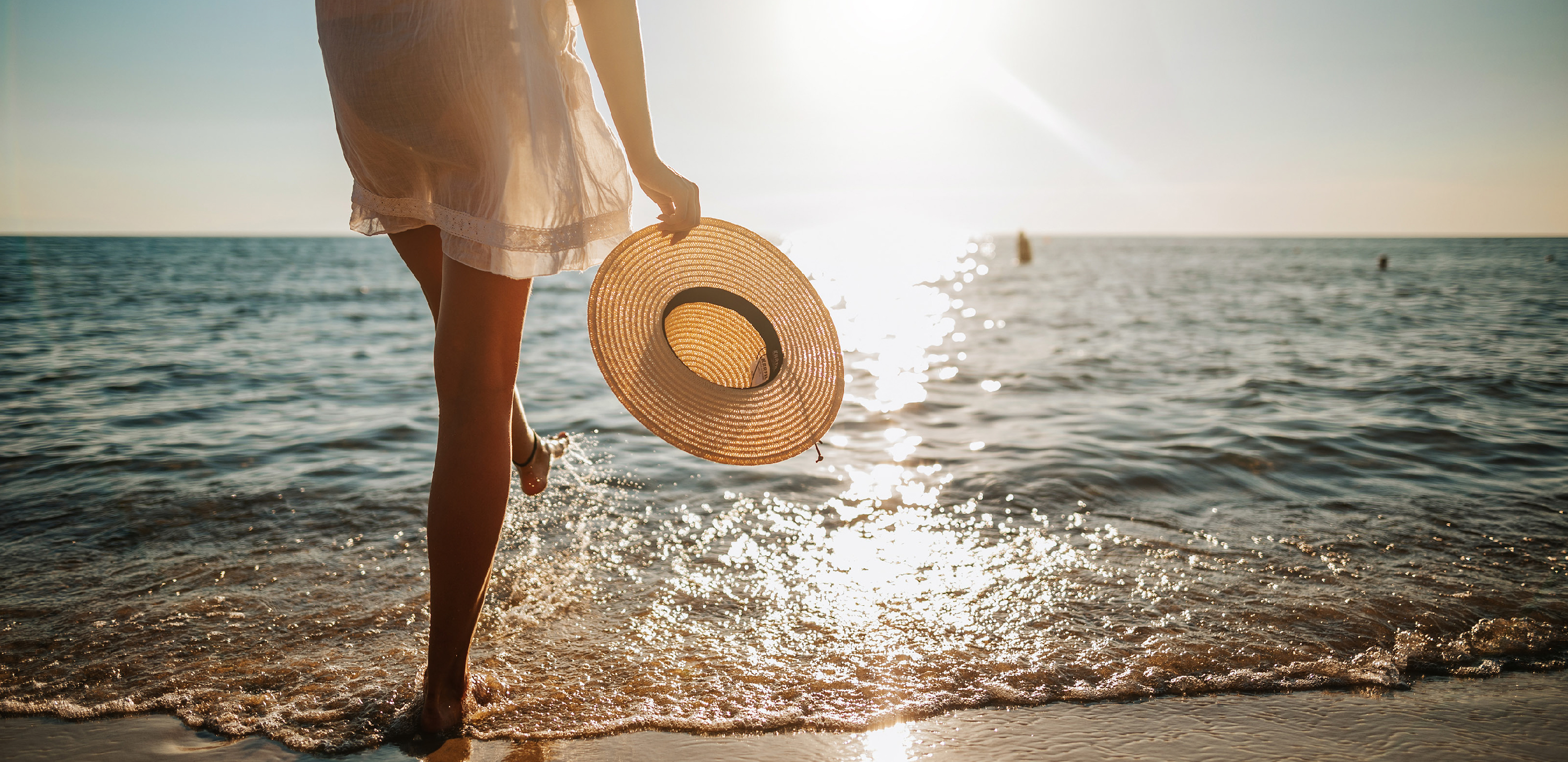 A woman walks along a sunny beach holding a sun hat