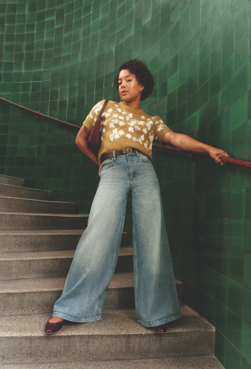 A model wears wide leg blue denim jeans and a green/yellow top with white flowers on it.