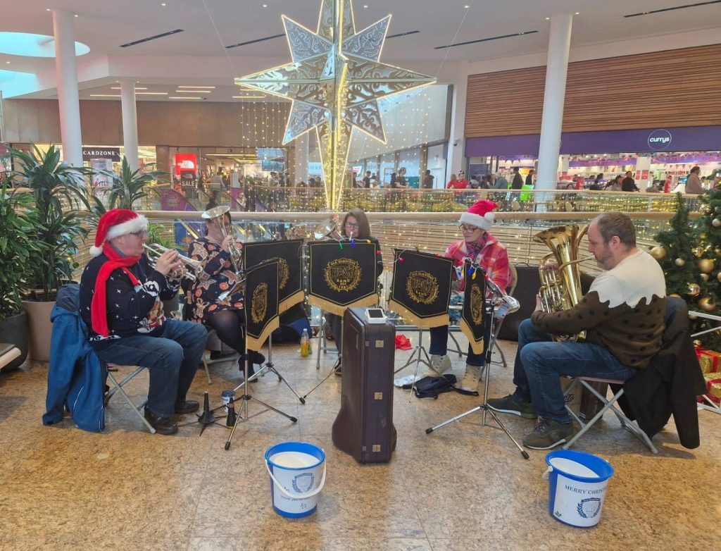 Five members of the brass band are shown playing instruments, including a tuba and cornets. They are wearing festive items like Santa hats, a Christmas jumper, and a scarf.
