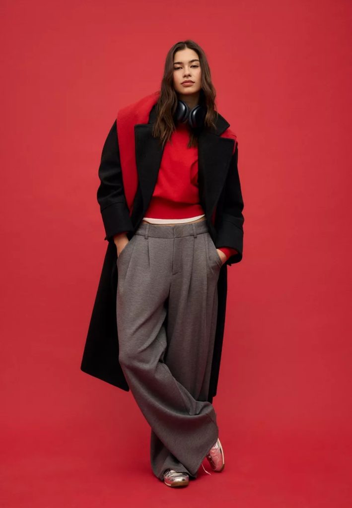 Model wears baggy grey trousers, a red jumper and a long black coat with red scarf. She is stood against a bright red background.