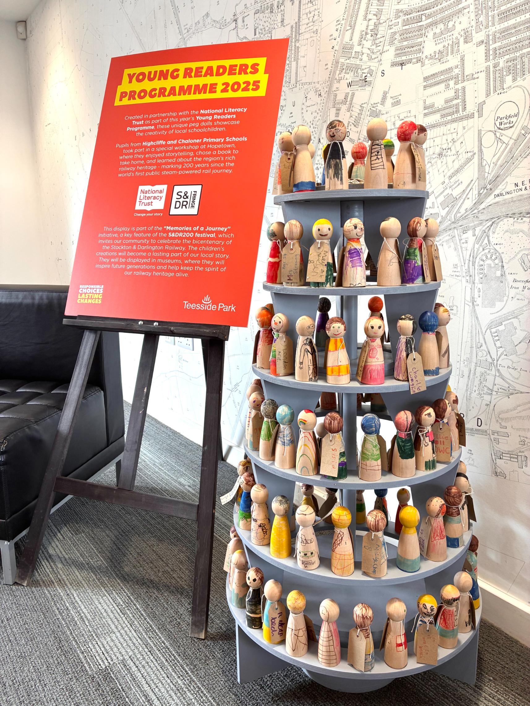 This image shows an indoor exhibition display, centered around a large, tiered sculpture made of painted wooden peg dolls. Each peg doll is uniquely decorated and many have small tags attached. The sculpture is a circular, multi-level stand, and it is positioned on the right side of the image.