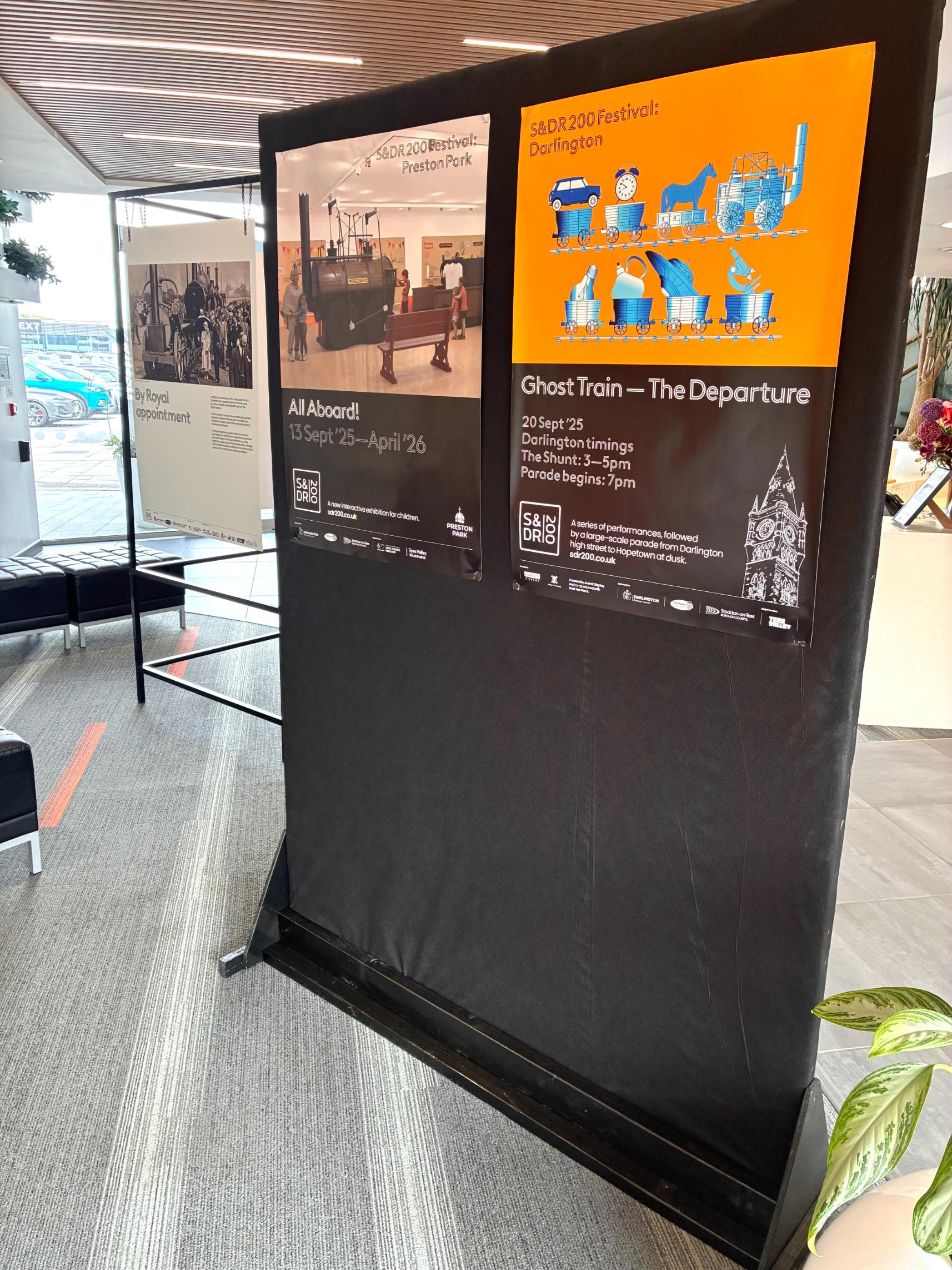 This image shows a two-sided exhibition display, likely in a public space like an office lobby or a museum. The display stand is covered in black fabric and holds two posters side-by-side on the side facing the camera.