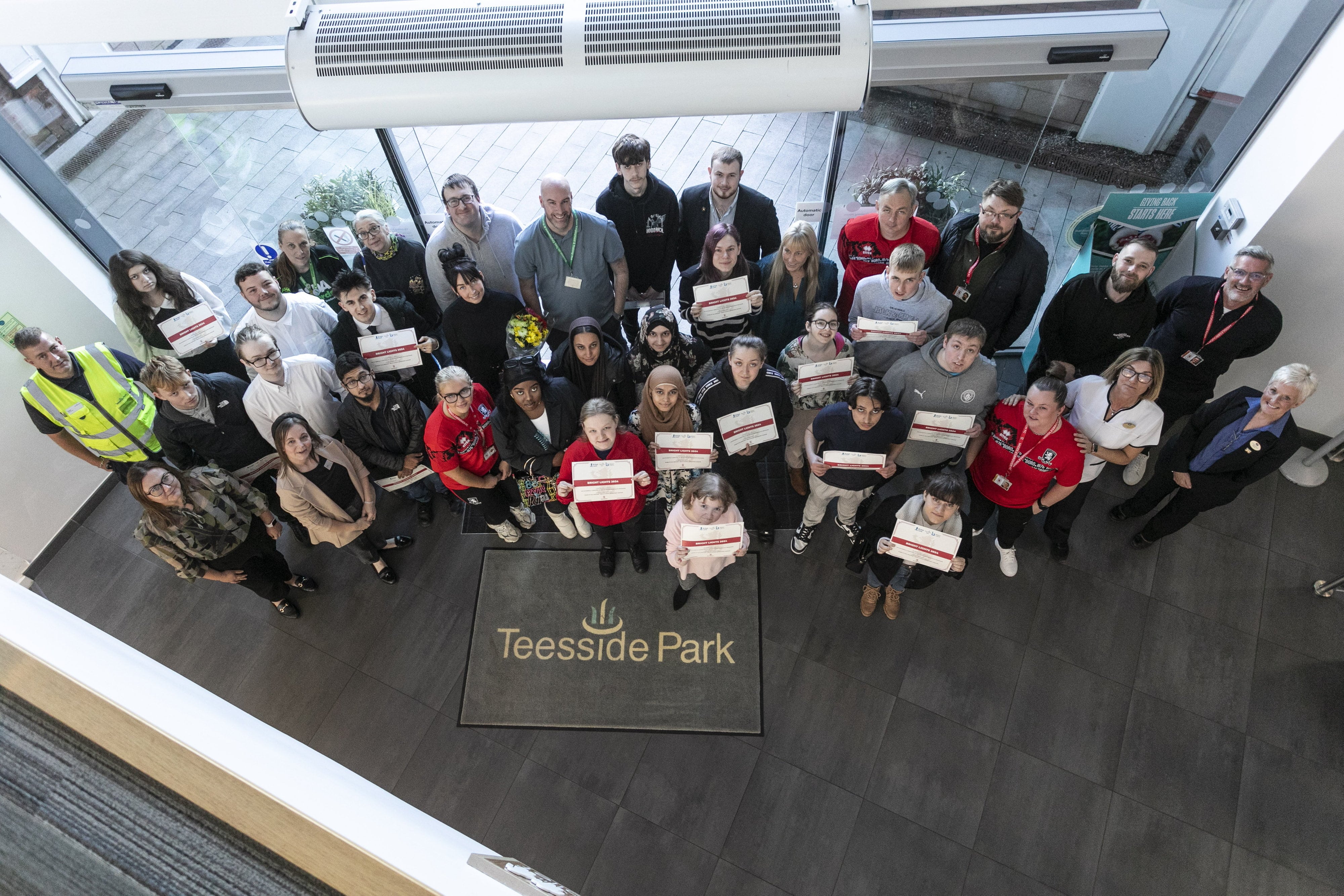 The photo likely captures a moment from an awards or graduation ceremony, as many individuals in the center of the group are holding certificates. The group is large and diverse, suggesting they are participants, staff, or partners from a community program, training course, or an initiative associated with the park. Some people are wearing uniforms or branded clothing, possibly representing the organizations involved.